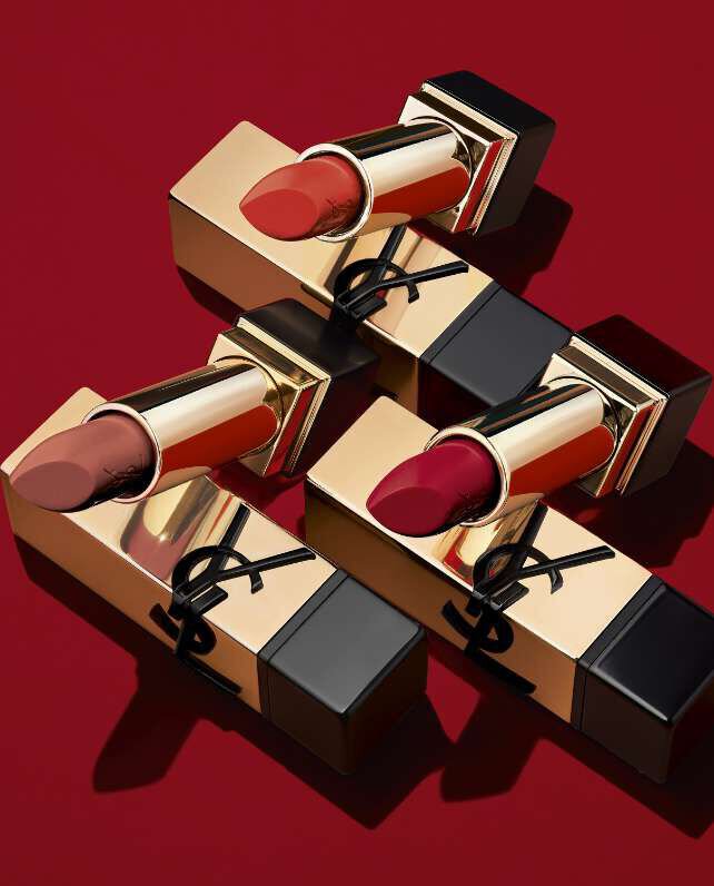 Close-up of three luxurious lipsticks in gold tubes, arranged on a rich red background, showcasing various shades of red and nude.