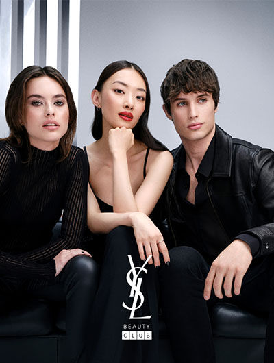 Three stylish models seated together on a black couch, showcasing modern fashion against a sleek gray backdrop. The central figure poses confidently with red lipstick, while the others wear elegant black outfits, representing a fashionable beauty campaign. The Yves Saint Laurent Beauty logo is prominently featured.
