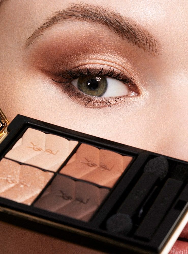 A close-up of a woman's eye with shimmery brown eyeshadow, holding a makeup palette featuring neutral shades.