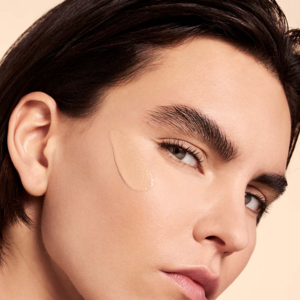 A close-up of a person with a smooth complexion, featuring a subtle application of makeup on their cheek. They have dark, styled hair and prominent brows, with a neutral background.