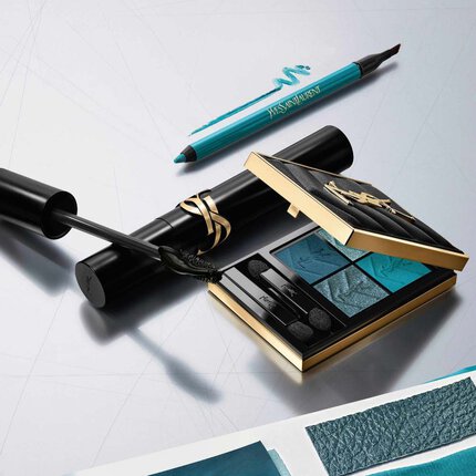 A luxury YSL beauty flat lay featuring an open gold-trimmed eyeshadow palette with blue and teal shades, a black mascara wand with product, and a teal eyeliner pencil, all arranged on a reflective silver surface.