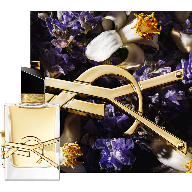 Libre Eau de Parfum Flakon, inszeniert mit goldener Kette und violetten Blumen vor dunklem Hintergrund.