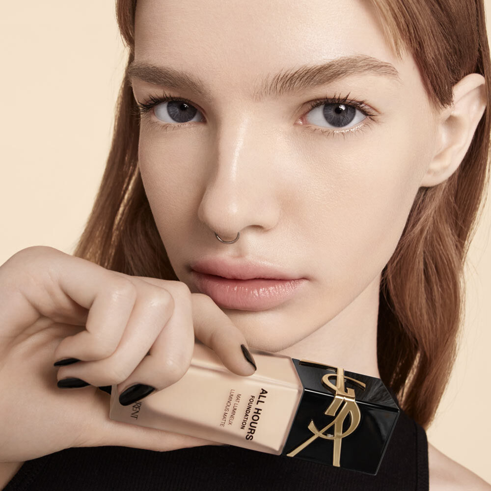 A close-up of a woman with fair skin and light brown hair, holding a bottle of foundation labeled "ALL HOURS." She has minimal makeup and a septum piercing, with a neutral background.