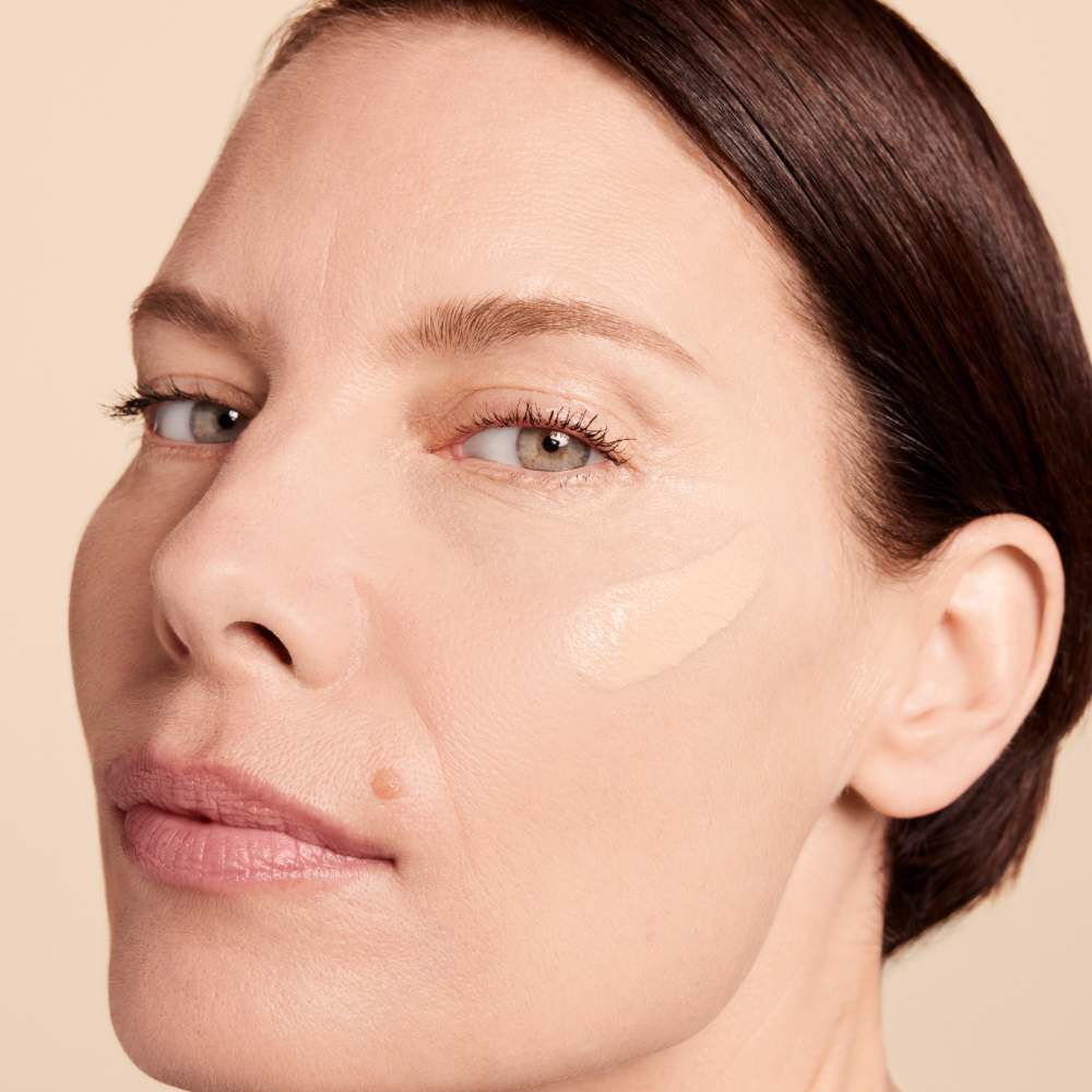 A close-up of a woman's face applying foundation to her cheek, with a neutral background. She has smooth skin and a slight smile.