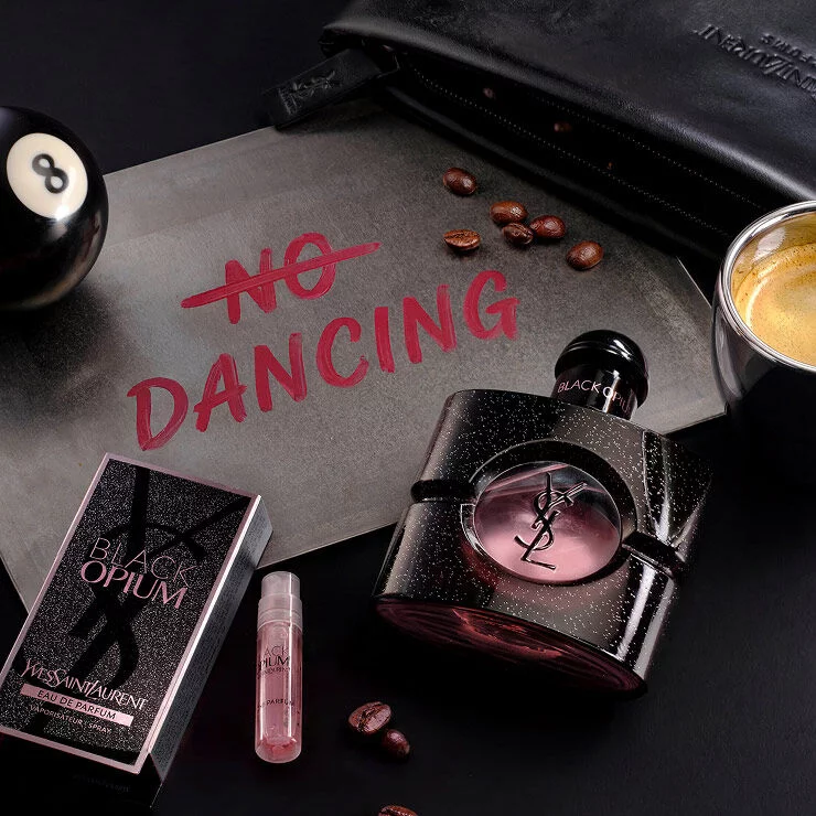 Black Opium Eau de Parfum and its sample vial next to a "No Dancing" sign, coffee beans, and a billiard ball.