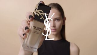 A model holds a bottle of YSL All Hours Foundation in front of a neutral background, showcasing its packaging.