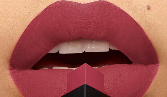 Close-up of a pair of lips adorned with a rich, matte lipstick color, holding a lipstick bullet at the center. The lips are slightly open, showcasing a natural tooth color.