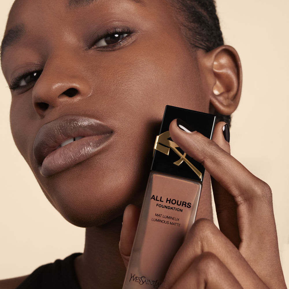 A close-up of a person holding a bottle of "All Hours Foundation" by Yves Saint Laurent, showcasing their smooth skin and neutral makeup look, with a soft background.