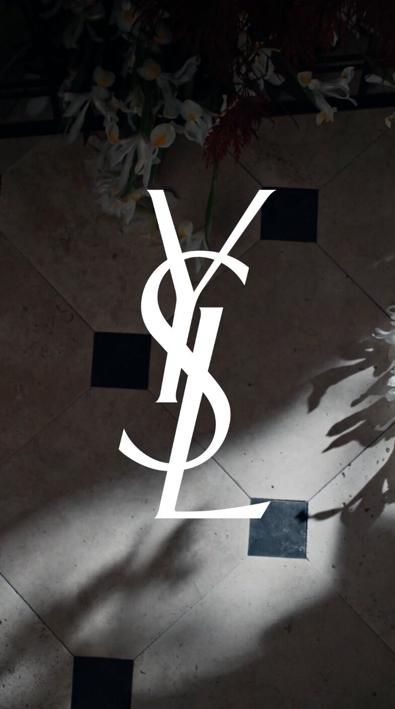 A floor featuring a geometric pattern with shadows cast by surrounding plants, overlaid with the iconic YSL logo in white.