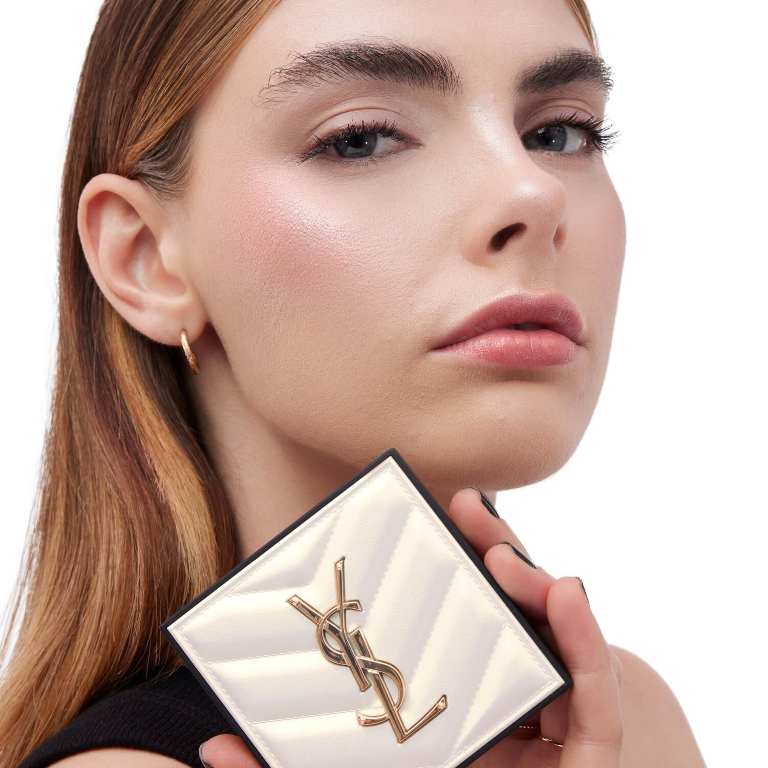 A close-up of a model holding a YSL cosmetic compact, with a clean and neutral expression, showcasing natural makeup and subtle highlights.