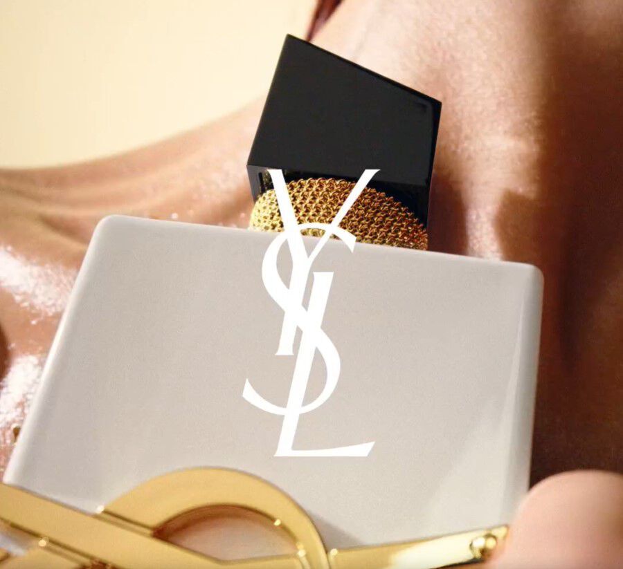 A close-up of a luxurious perfume bottle with a black and gold cap, featuring the YSL logo in white on a matte surface, partially resting against a skin-like background.