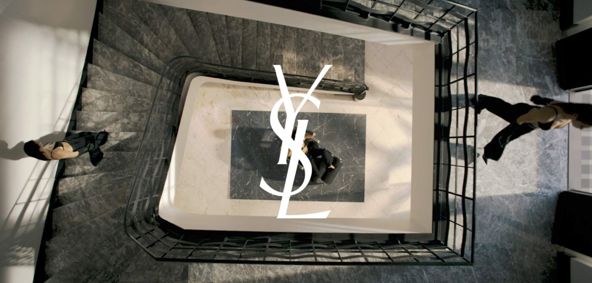 Aerial view of a black and white staircase with two figures walking on different levels, featuring the YSL logo prominently in the center.