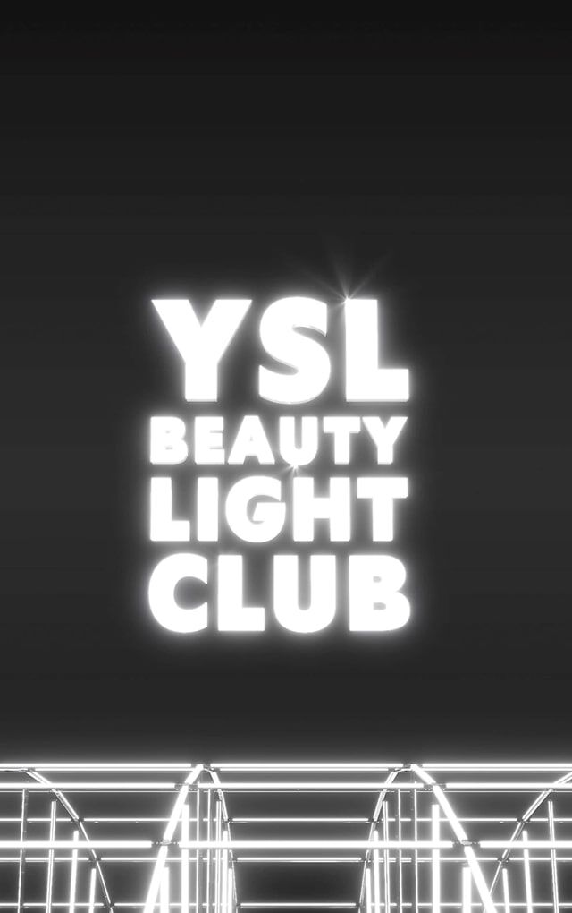Eine weite, dunkle Aufnahme zeigt das Logo „YSL BEAUTY LIGHT CLUB“ in leuchtend weißen Buchstaben, schwebend in der Mitte eines schwarzen Raums. Unterhalb des Logos erzeugt eine komplexe Struktur aus Metallstangen oder leuchtenden weißen Neonlichtern einen Gitter- oder Buhneneffekt, der eine Lichtinstallation oder eine futuristische Clubumgebung suggeriert. Der Gesamteindruck ist der einer modernen und schicken Atmosphare.