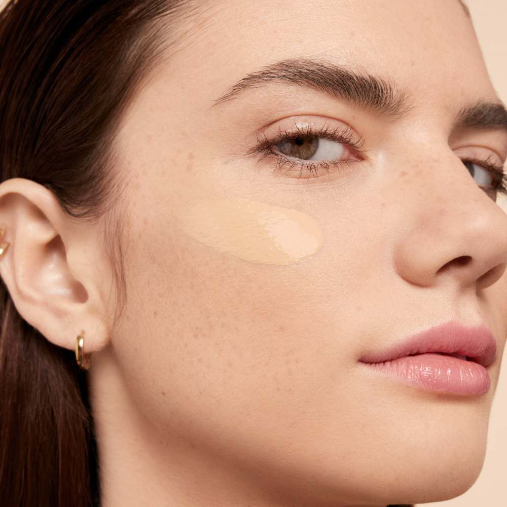 Close-up of a woman applying foundation on her cheek, showcasing her smooth skin and natural freckles.