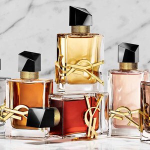 Eine Hand halt einen roten YSL-Parfumflakon neben zwei weiteren Duften auf einem Steinsockel.