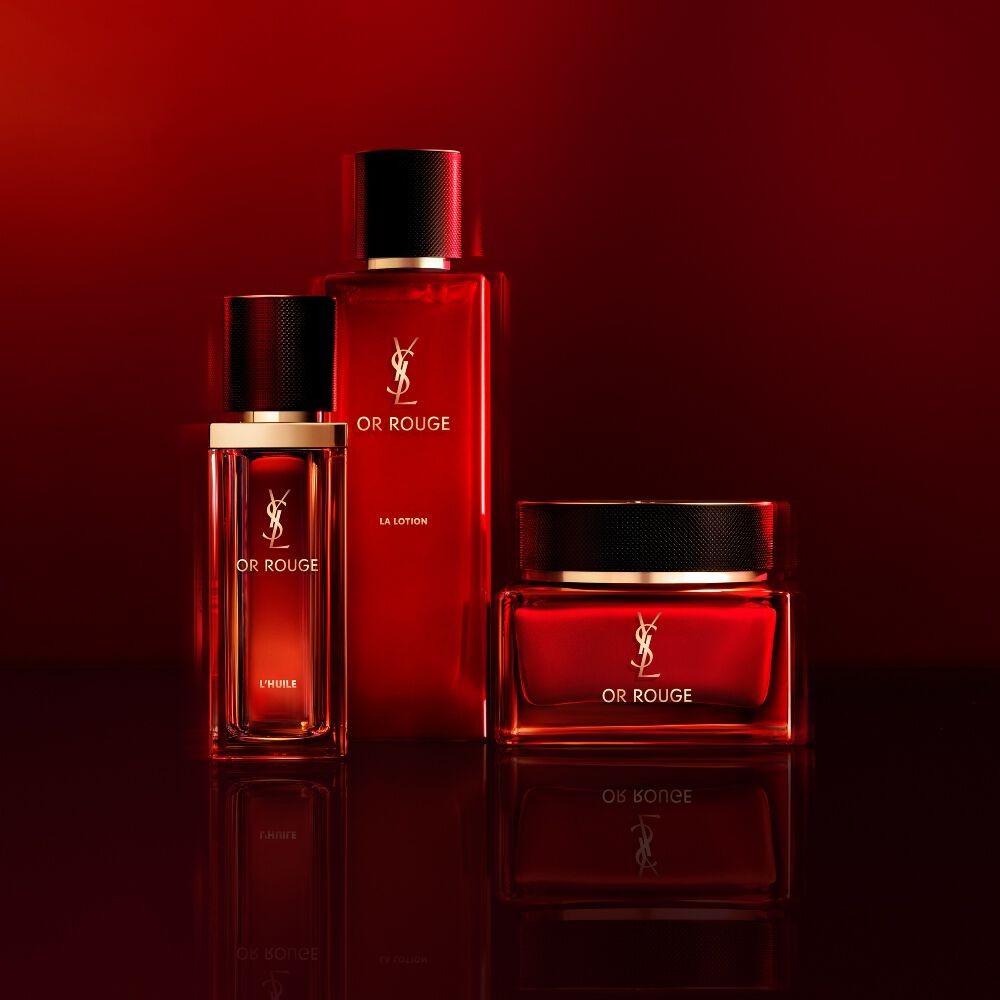 Drei Hautpflegeprodukte von Yves Saint Laurent Or Rouge in lichtdurchlassigen roten Glasflaschen und einem Tiegel auf dunkelrotem Hintergrund.