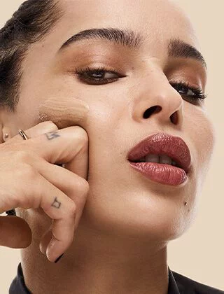 A close-up portrait of a woman applying foundation to her face with a finger, showcasing a confident expression and glossy lips against a light beige background.