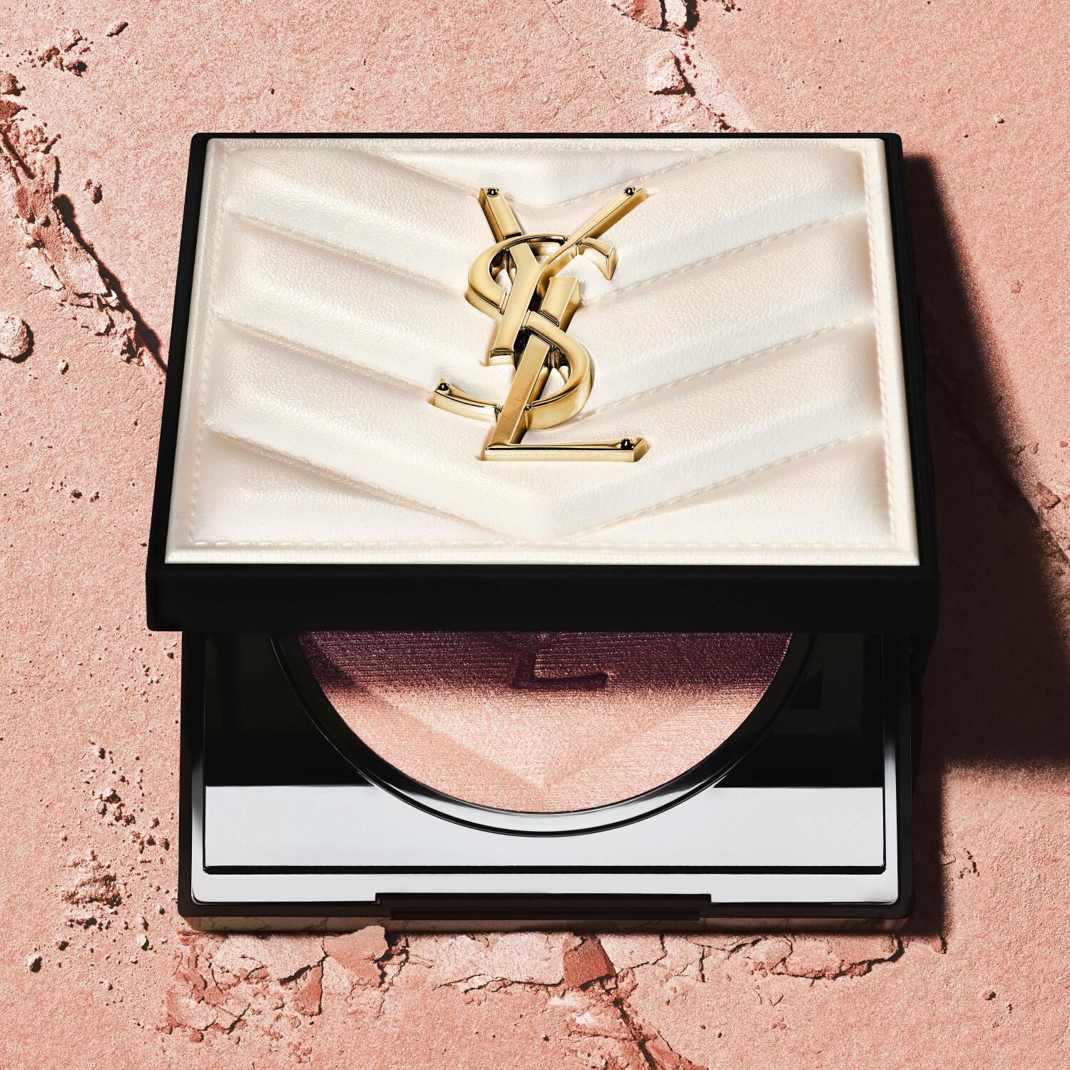 Alt text: A close-up of a YSL beauty product compact with a chevron-patterned lid, featuring the YSL logo, resting on a textured pink surface.