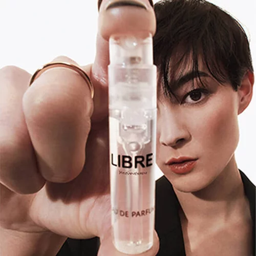 Eine Person mit kurzen dunklen Haaren halt ein kleines, durchsichtiges Parfumprobenflaschchen mit der Aufschrift „LIBRE Yves Saint Laurent Eau de Parfum“ hoch. Ihr Gesicht, mit direktem Blick, ist teilweise hinter ihrer Hand und dem Flaschchen zu sehen.