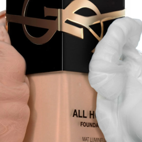 A bottle of foundation labeled "All Hours" by Yves Saint Laurent, held between two hands, one gloved in white and the other bare.