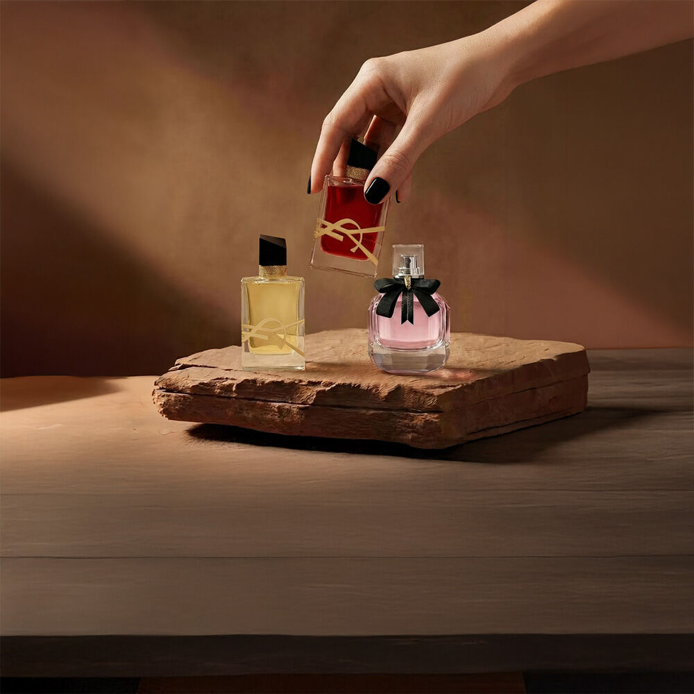 Eine Hand halt einen roten YSL-Parfumflakon neben zwei weiteren Duften auf einem Steinsockel.