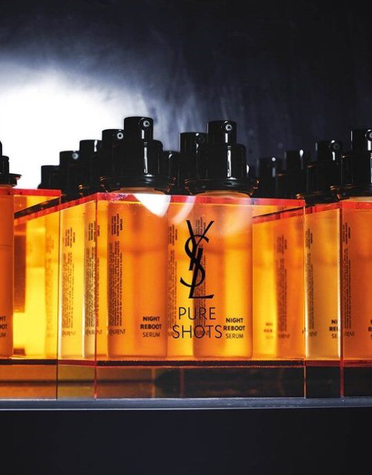 Reihe bernsteinfarbener Yves Saint Laurent Pure Shots Night Reboot Serumflaschen mit schwarzen Pumpverschlussen. Das YSL-Logo ist auf jeder Flasche deutlich sichtbar, die auf einer reflektierenden Oberflache vor einem dunklen, rauchigen Hintergrund steht und die Produktlinie hervorhebt.