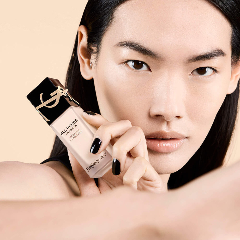 A close-up of a model holding a bottle of YSL All Hours foundation, showcasing flawless skin and dark hair against a soft beige background. The model has a neutral expression and striking nails.