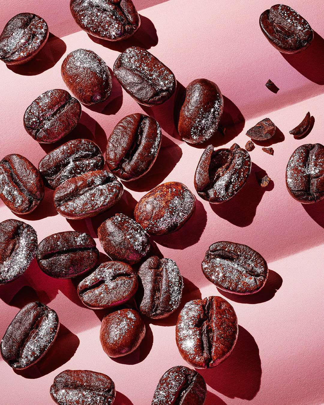 A close-up of scattered dark roasted coffee beans dusted with a light coating of powdered sugar on a smooth pink surface.