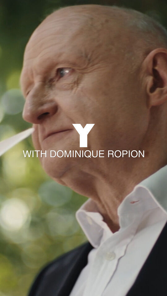 Ein Mann mit Glatze, schwarzem Anzug und weißem Hemd halt sich mehrere weiße Papierstreifen, die ublicherweise fur Parfumtests verwendet werden, vor die Nase, als ob er einen Duft bewerten wurde. Er sieht nachdenklich aus, mit einem grunen, verschwommenen Hintergrund aus Laub und Baumen. Uber dem Bild steht in großer weißer Schrift "Y" und darunter in kleinerer Schrift "WITH DOMINIQUE ROPION".