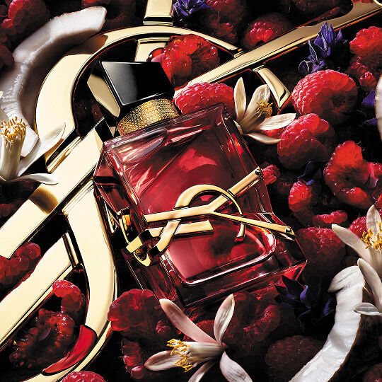 Der rosa Flakon von Yves Saint Laurent Libre Berry Crush, umgeben von Himbeeren, weißen Orangenbluten und Kokosnussstucken auf einem goldenen Logo-Hintergrund.