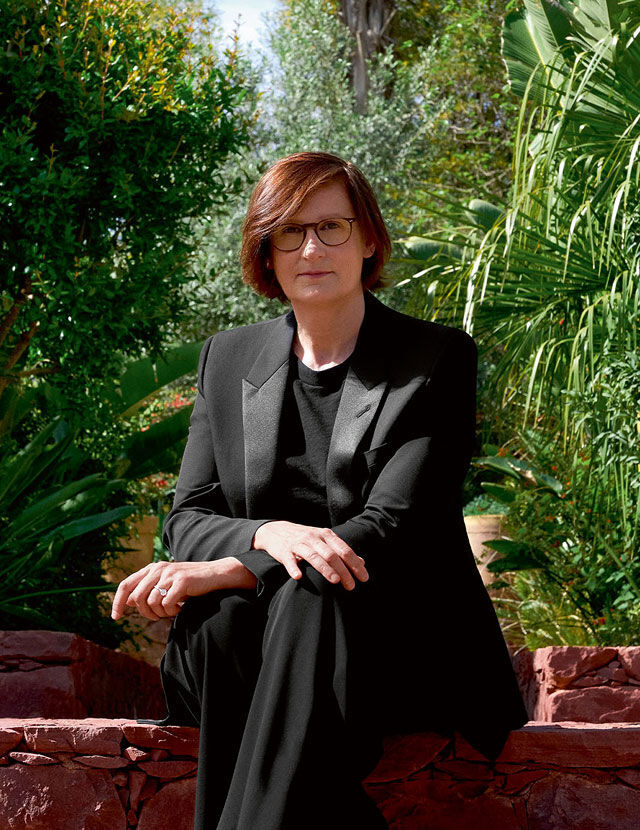 Eine Frau mit rotlich-braunem Haar und Brille sitzt auf einer Steinmauer und tragt einen schwarzen Anzug. Sie schaut den Betrachter an, mit einem uppigen grunen Garten im Hintergrund.