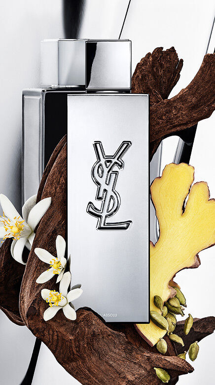 YSL Y L'ABSOLU Parfumflakon umgeben von dunklem Holz, weißen Orangenbluten, Ingwer und Kardamomkapseln.