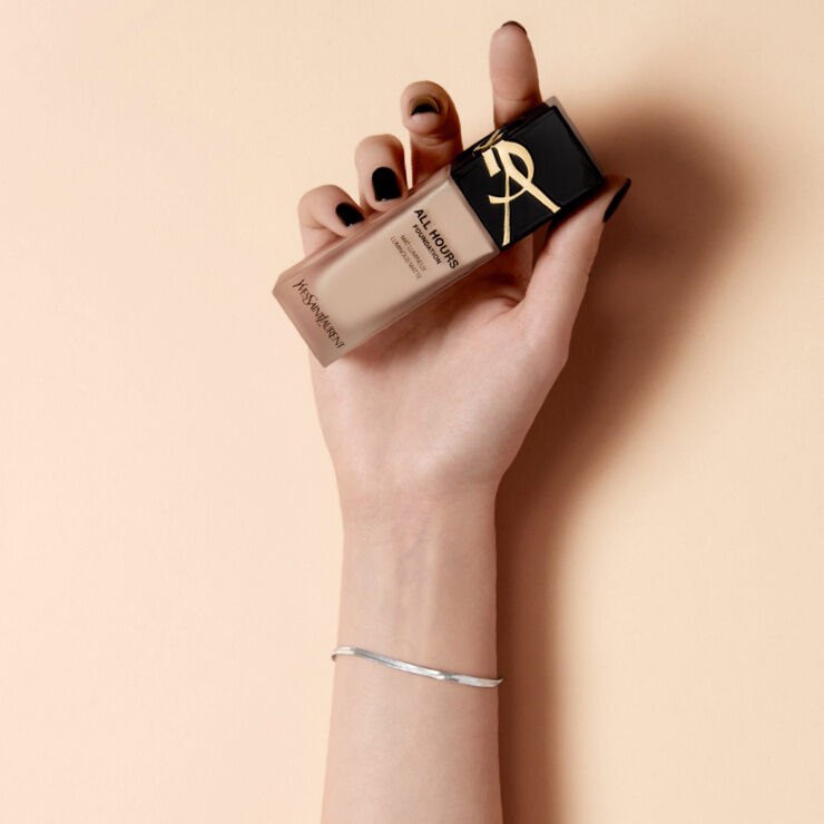 A hand with black nail polish holding a bottle of YSL foundation against a soft beige background.