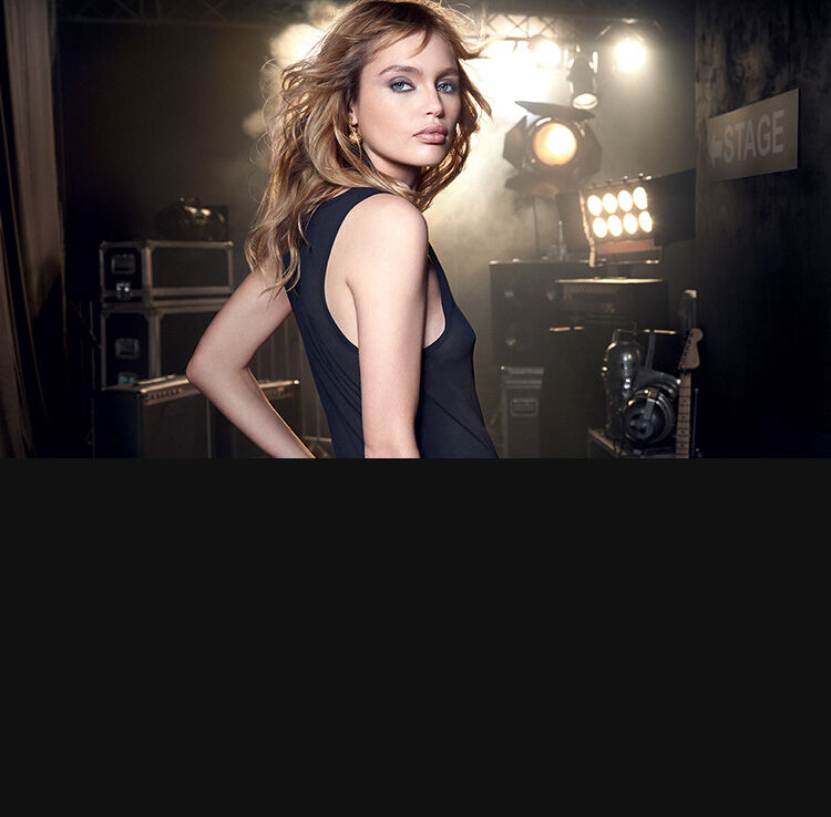 A stylish model poses confidently with long wavy hair, wearing a sleek black outfit. The background features stage lights and musical equipment, suggesting a backstage setting.