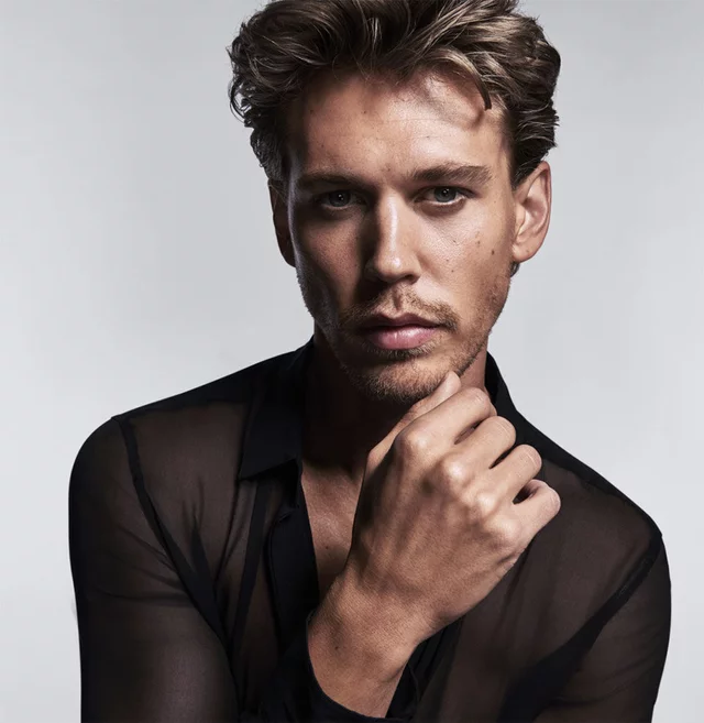 A close-up portrait of a young man with tousled hair, wearing a sheer black shirt. He is looking directly at the camera with a serious expression, resting his chin on his hand. The background is a soft gradient of light gray.