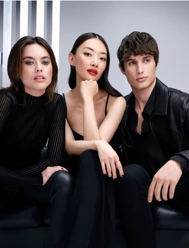 Drei stilvolle junge Models in schwarzer Kleidung sitzen auf einem schwarzen Leder-Sofa vor einem grauen Studio-Hintergrund.