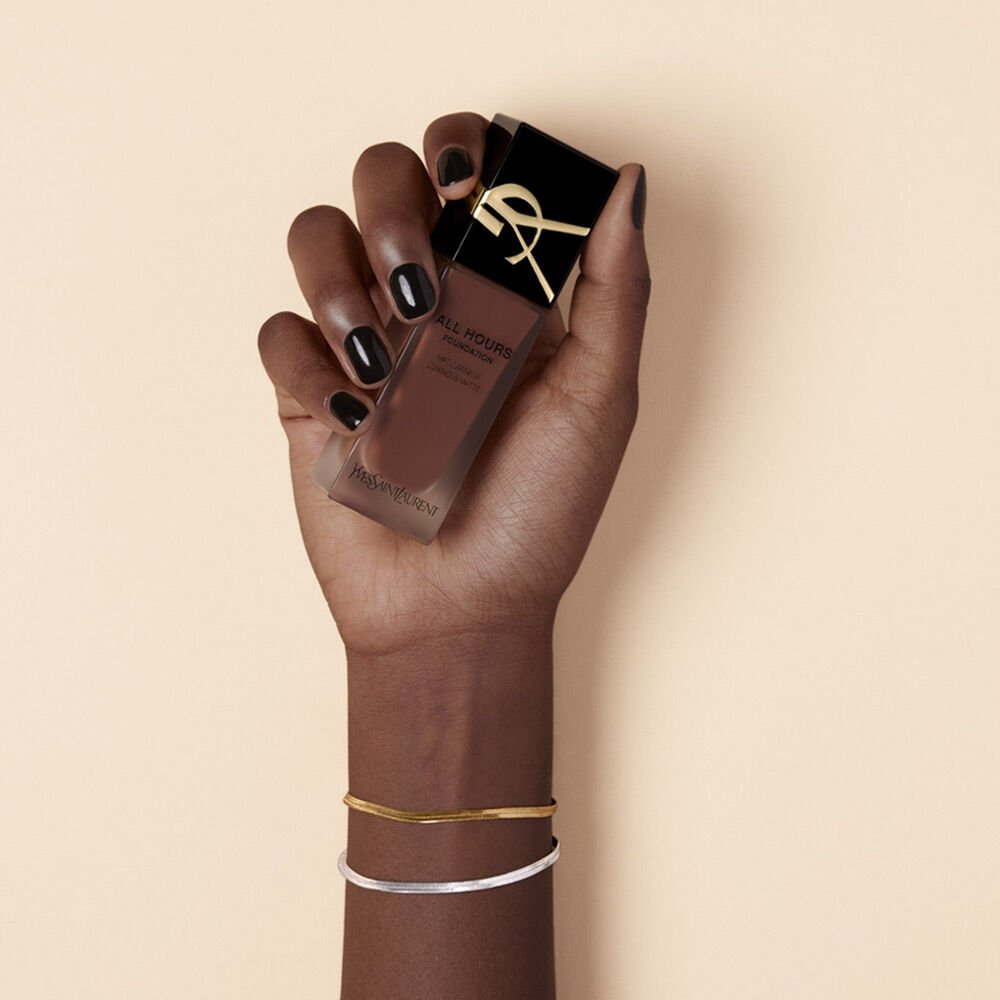 A hand with dark nail polish holds a bottle of YSL All Hours foundation against a neutral background.