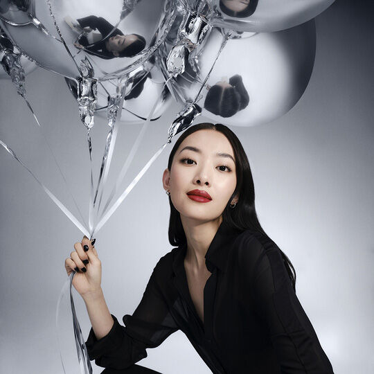 Eine Frau mit langen dunklen Haaren, die ein schwarzes Hemd tragt, halt silberne Luftballons und schaut direkt in die Kamera. Sie tragt roten Lippenstift.