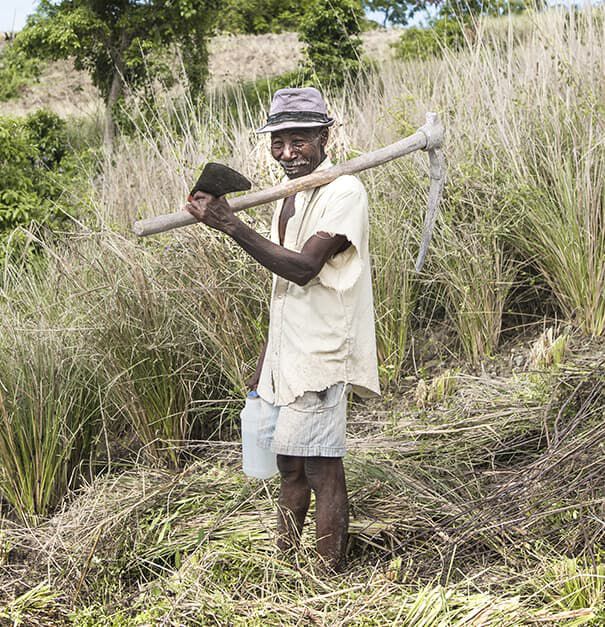 A farmer standing in a grassy field, holding a pickaxe over his shoulder and wearing a hat and a light shirt. He has a water container next to him and is smiling.