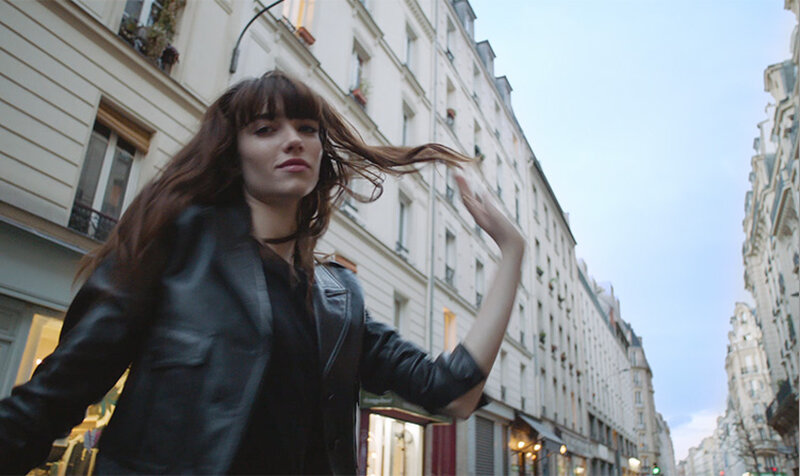 A young woman with long brown hair is dancing in a charming European street lined with buildings. She wears a black leather jacket and has a confident expression as her hair flows in the breeze.