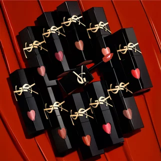 A collection of stylish black lipstick tubes with gold logos, arranged in a circular pattern on a vibrant red surface. Some tubes have heart-shaped tips showing various shades of lipstick inside.