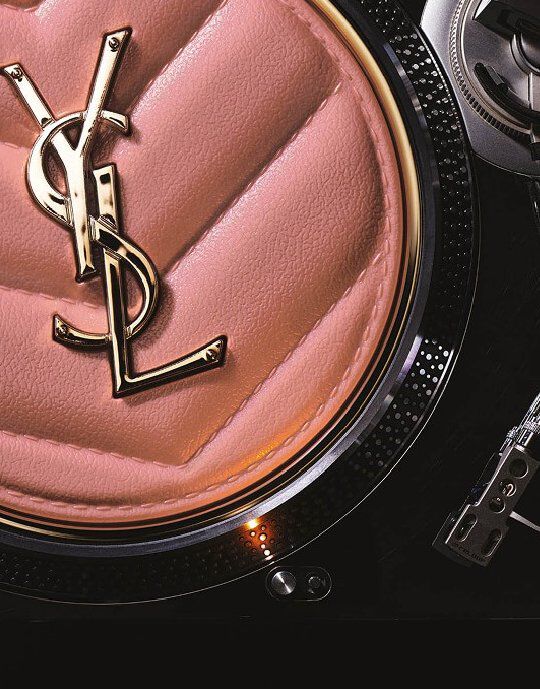 Nahaufnahme eines rosa, gesteppten Yves Saint Laurent Puderdosen oder Accessoires mit einem goldenen YSL-Logo in der Mitte. Der Gegenstand liegt auf dem metallischen, strukturierten Rand eines Plattenspielers, was Luxus-Beauty mit einer musikalisch inspirierten Asthetik verbindet.