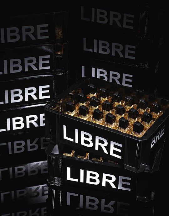 Eine Komposition aus schwarzen Kisten, ahnlich denen fur den Flaschentransport, gestapelt und gefullt mit Flakons des YSL Libre Parfums. Die Kisten tragen die Aufschrift „LIBRE“ in großen, leuchtend weißen Buchstaben. Im Vordergrund ist eine Kiste mit zahlreichen „Libre“-Parfumflakons gefullt, die das Licht reflektieren und goldene Glanzpunkte erzeugen. Das gesamte Arrangement ist in ein gedampftes Licht getaucht, was den schicken Charakter und die Inszenierung des Produkts betont.