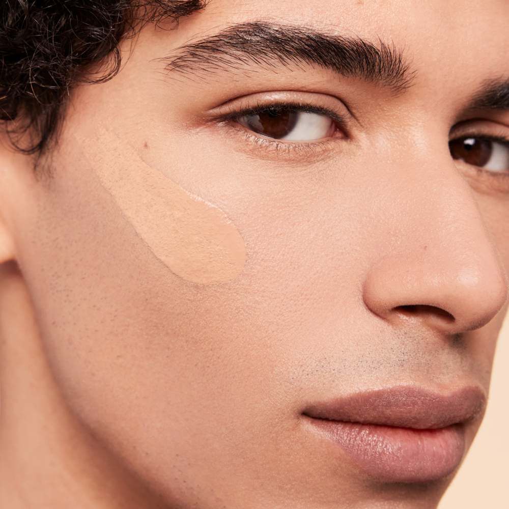 A close-up of a young man with a light complexion, showcasing a swatch of foundation applied on his cheek. His expression is neutral, and he has curly hair.