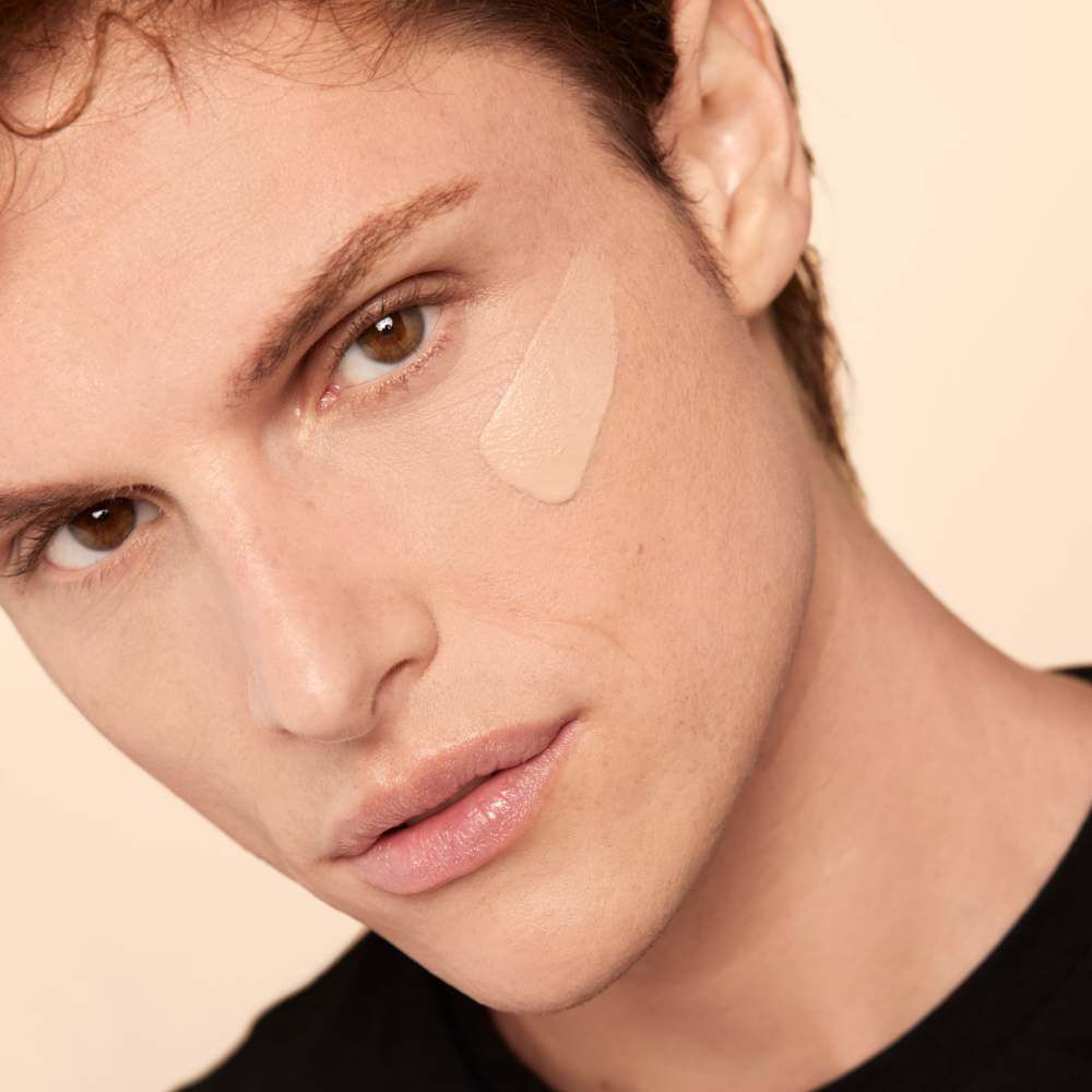A close-up of a young man applying foundation on his cheek, with a focused expression and warm lighting.