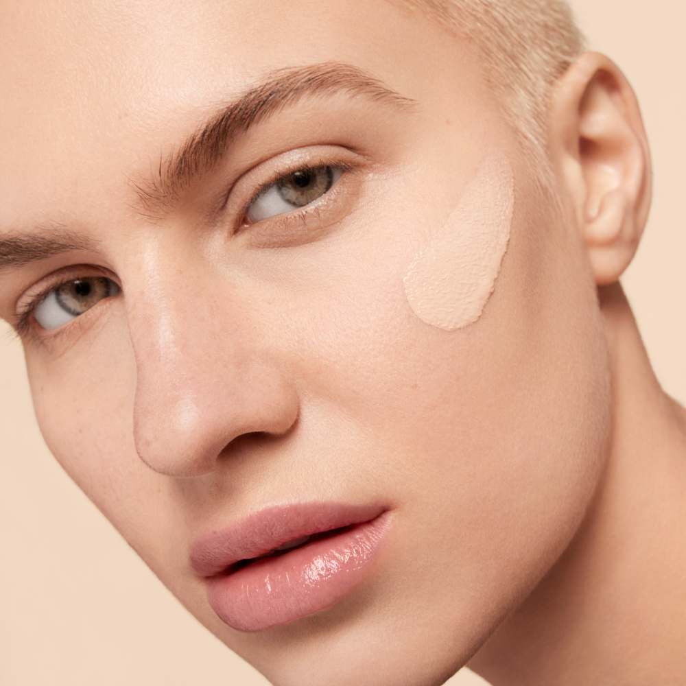 A close-up of a person with short, light-colored hair, applying a foundation or concealer to their cheek, revealing smooth skin and neutral makeup tones.