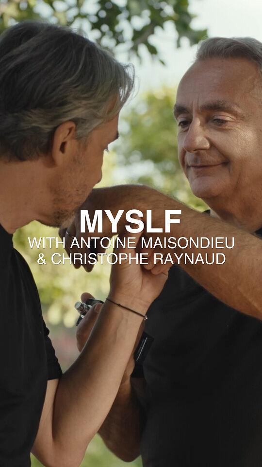 Zwei Manner in schwarzen T-Shirts interagieren im Freien, wobei einer den Arm des anderen und einen kleinen Gegenstand halt. Das Bild ist mit dem Text "MYSLF WITH ANTOINE MAISONDIEU & CHRISTOPHE RAYNAUD" vor einem verschwommenen grunen Laubhintergrund uberlagert.