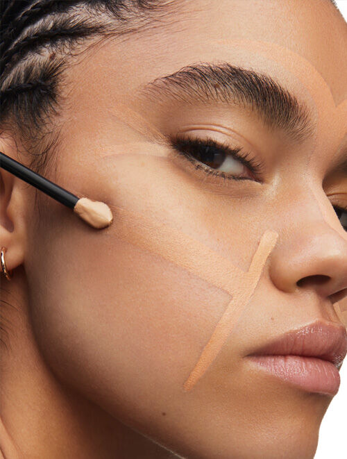 A close-up of a woman's face applying foundation with a brush, featuring defined strokes of makeup on her skin. She has braided hair and a natural, neutral expression.