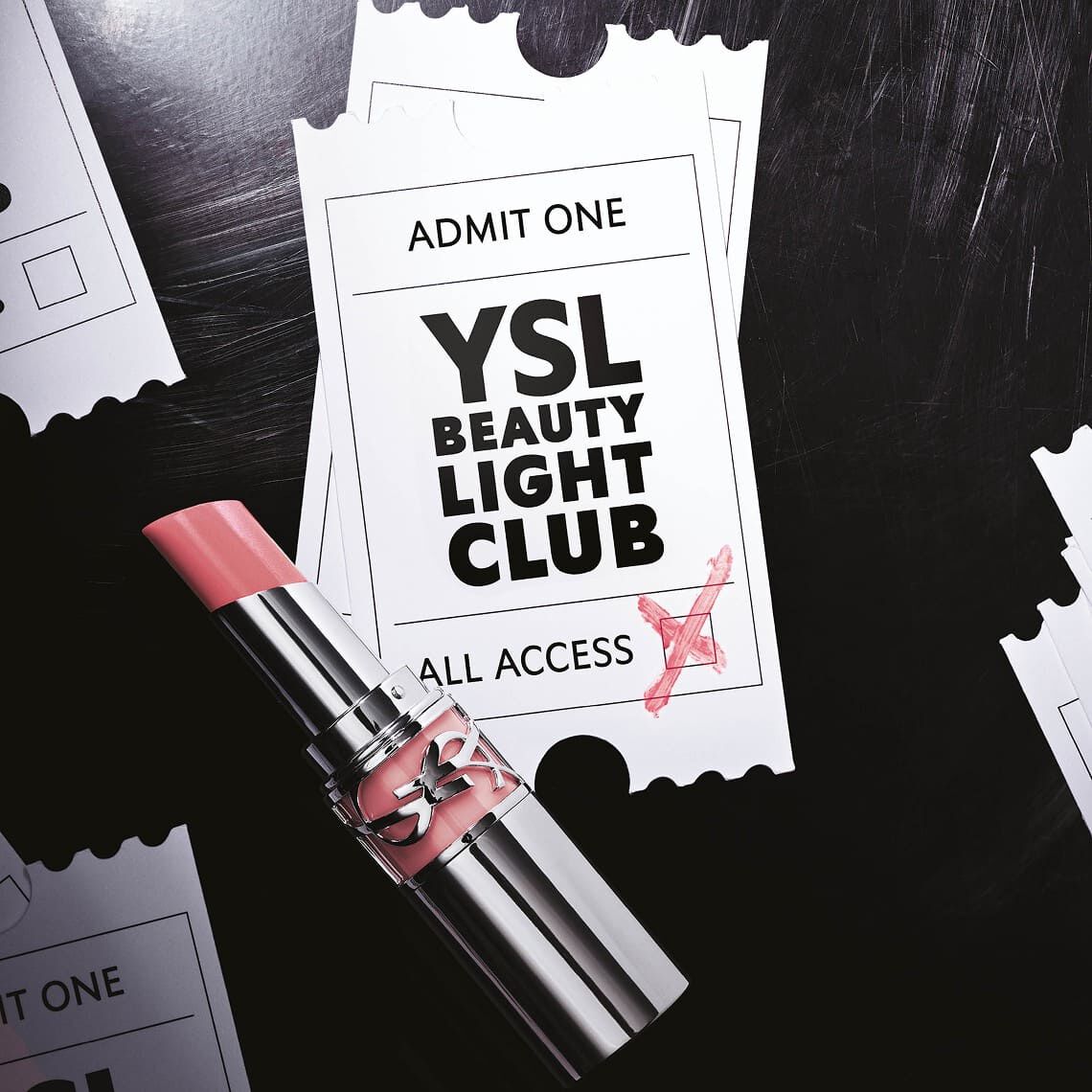 Ein hellrosa Lippenstift mit glanzender Textur liegt auf zwei leicht uberlappenden weißen Event-Tickets. Das obere Ticket zeigt oben den Text „ADMIT ONE“ und in der Mitte in großen schwarzen Buchstaben „YSL BEAUTY LIGHT CLUB“, darunter „ALL ACCESS“. Ein rotes Kreuz ist auf die untere rechte Ecke des Tickets gekritzelt, als ob es validiert wurde. Der Hintergrund ist dunkel und erinnert an eine texturierte Oberflache, und an den Seiten sind zerfetzte Ticketrander sichtbar, die auf mehrere andere verstreute Tickets hinweisen. Der Gesamteindruck ist der einer exklusiven Einladung zu einem glamourosen Event.