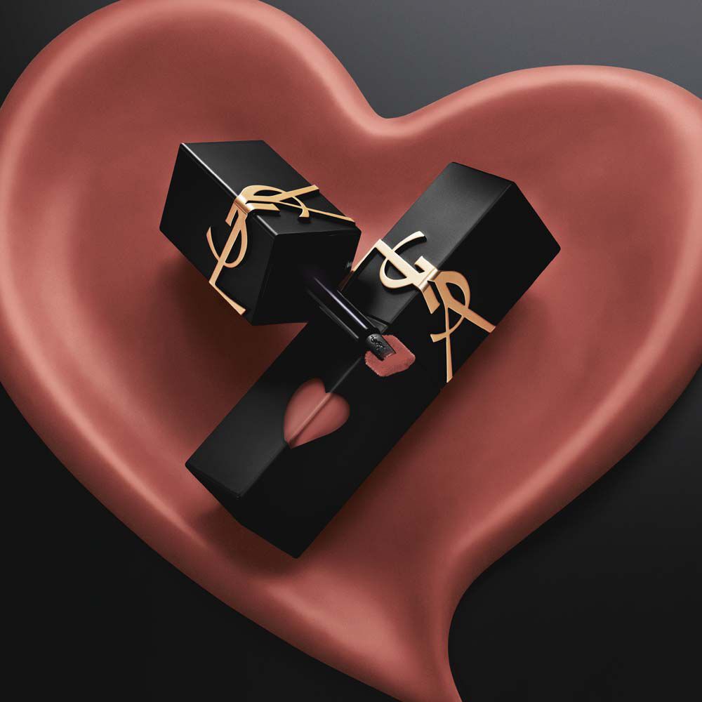 Zwei matte schwarze YSL-Lip-Stain-Flaschen mit pinkfarbenen Herzausschnitten und goldenen Logos – inszeniert in einem skulpturalen, matten pinken Herz auf schwarzem Grund.